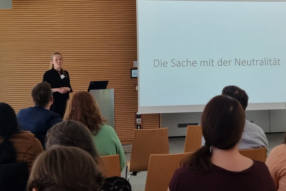 Prof. Dr. Inken Heldt hält einen Vortrag vor Publikum und auf der Powerpoint rechts neben ihr, steht geschrieben: Die Sache mit der Neutralität