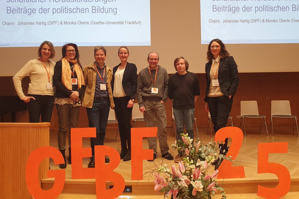 Prof. Dr. Inken Heldt steht in der Mitte auf einer Bühne mit Diskutant:innen, alle posieren für das Gruppenfoto. Im Vordergrund sind in roten aus Karton ausgeschnittene Buchstaben: GEBF 2025.