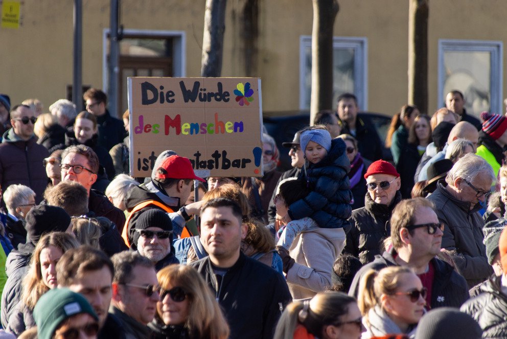 Eine Demonstration mit Menschen auf denen ein Schild hochgehalten wird, worauf zu lesen ist: "Die Würde des Menschen ist unantastbar"